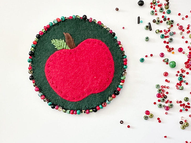 Felt apple pin on surface next to scattered, multi-colored beads