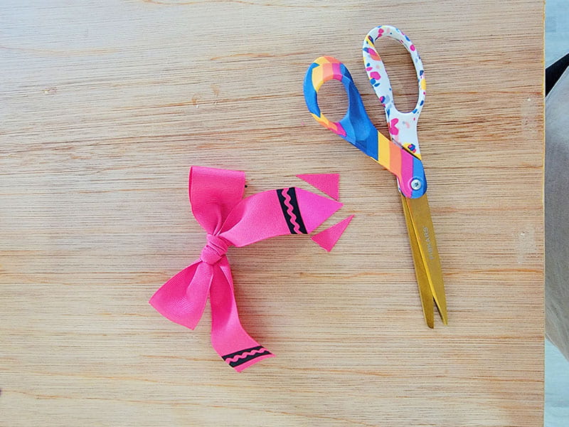 Pink crayon hair bow with fabric cut into triangle on right side with scissors next to it. 