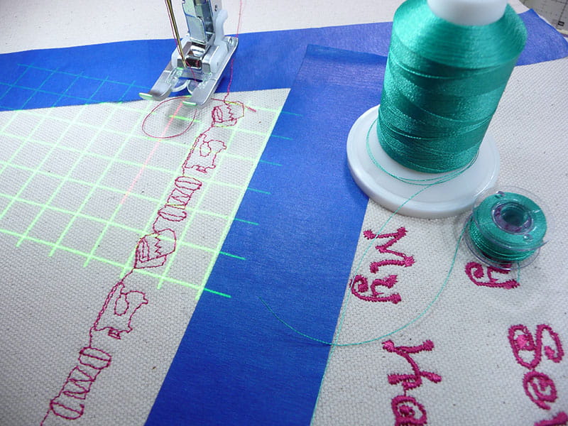 Teal thread on fabric cover. Embroidery machine foot visible next to a design in pink and laser light grid below.