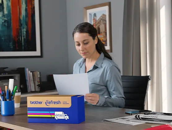 Woman sitting at a desk reviewing a document with a Refresh subscription box for toner at the front of the desk.  