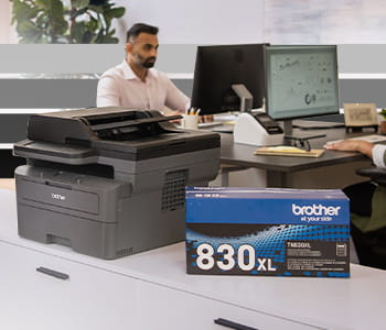 Brother ELLe black and white printer next to a TN830XL toner box with people sitting at desks in background.