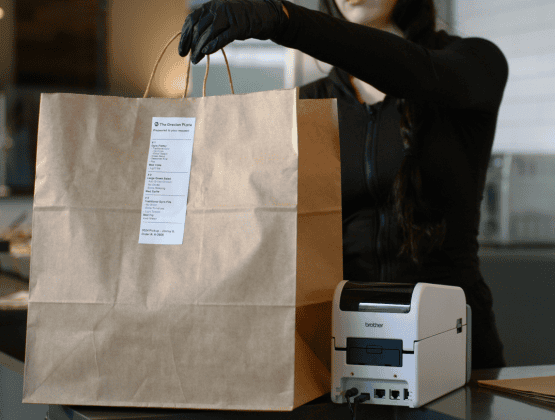 A gloved hand places a brown paper bag with a printed receipt label onto a counter next to a small label printer, representing curbside pickup.