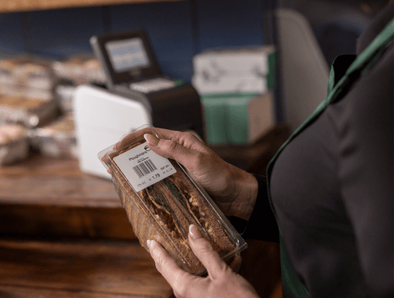 A person holds packaged bread while applying a printed label in a bakery or grocery store setting, illustrating fresh food labeling.