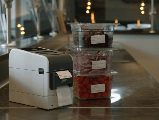 A label printer on a counter next to clear plastic containers filled with cut fruit, showing prepared food labeling.