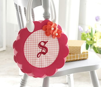 Pink flower bag with cursive S on gingham print background hanging on a white wooden chair