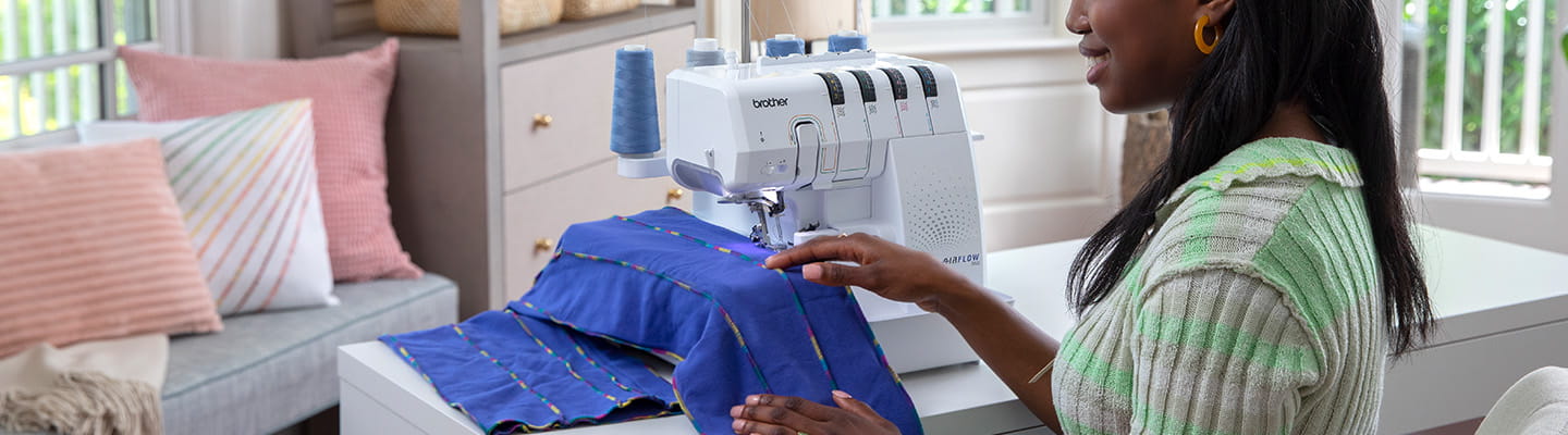Woman sitting at craft table using a Brother serger on blue fabric. 