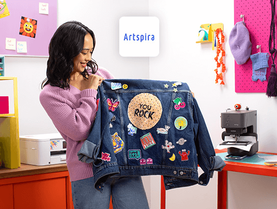 Woman holding jacket with patches designed with Artspira. Skitch embroidery machine and Brother sublimation printer in background