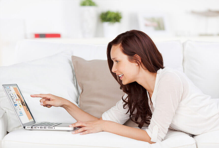 Woman lying on couch, pointing and laughing at laptop screen 