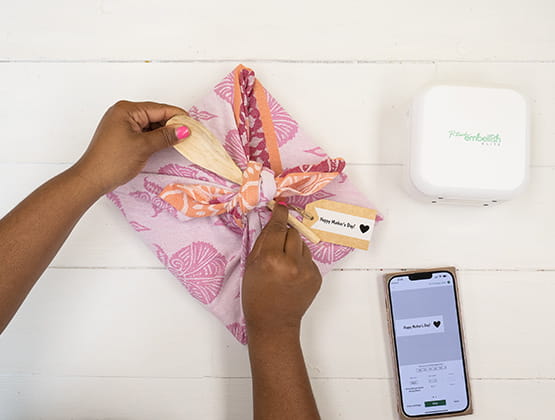 P-touch embellish craft step 3: Person adding P-touch Embellish ribbon and gift tag to finished gift. P-touch Label maker and cell phone next to gift.