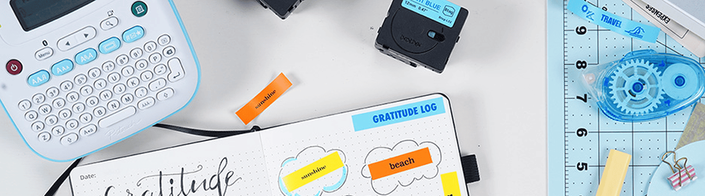 A flat lay of a Brother label maker, colorful label tape, ruler, and open gratitude journal showing labeled words like "beach" and "sunshine."