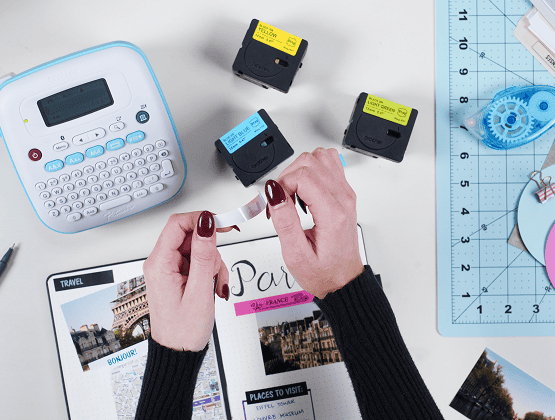 Hands unsticking the tape on a label maker near colorful tapes, preparing labels for a travel journal.