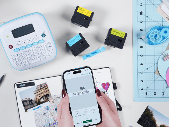 A phone displaying a print screen is held above a journal spread titled "Paris" with printed photo labels.