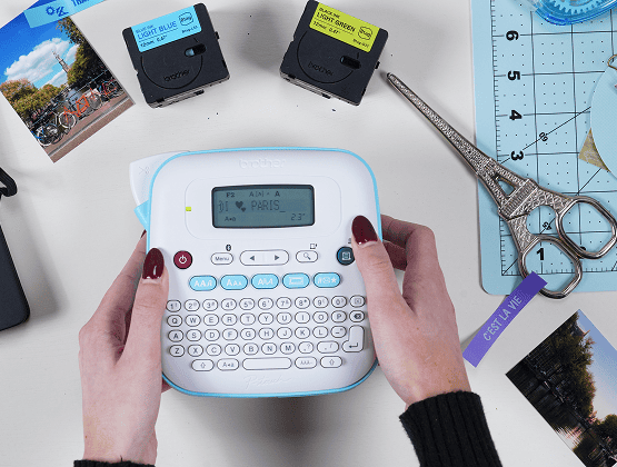 Hands typing on a label maker near printed photos, tapes, and crafting tools.