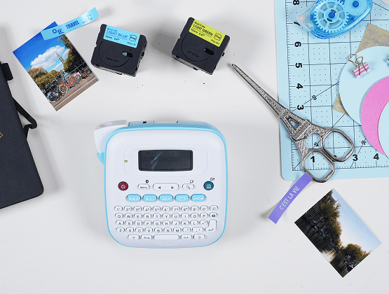 A white label maker surrounded by printed photos, scissors, and label tape on a crafting desk.
